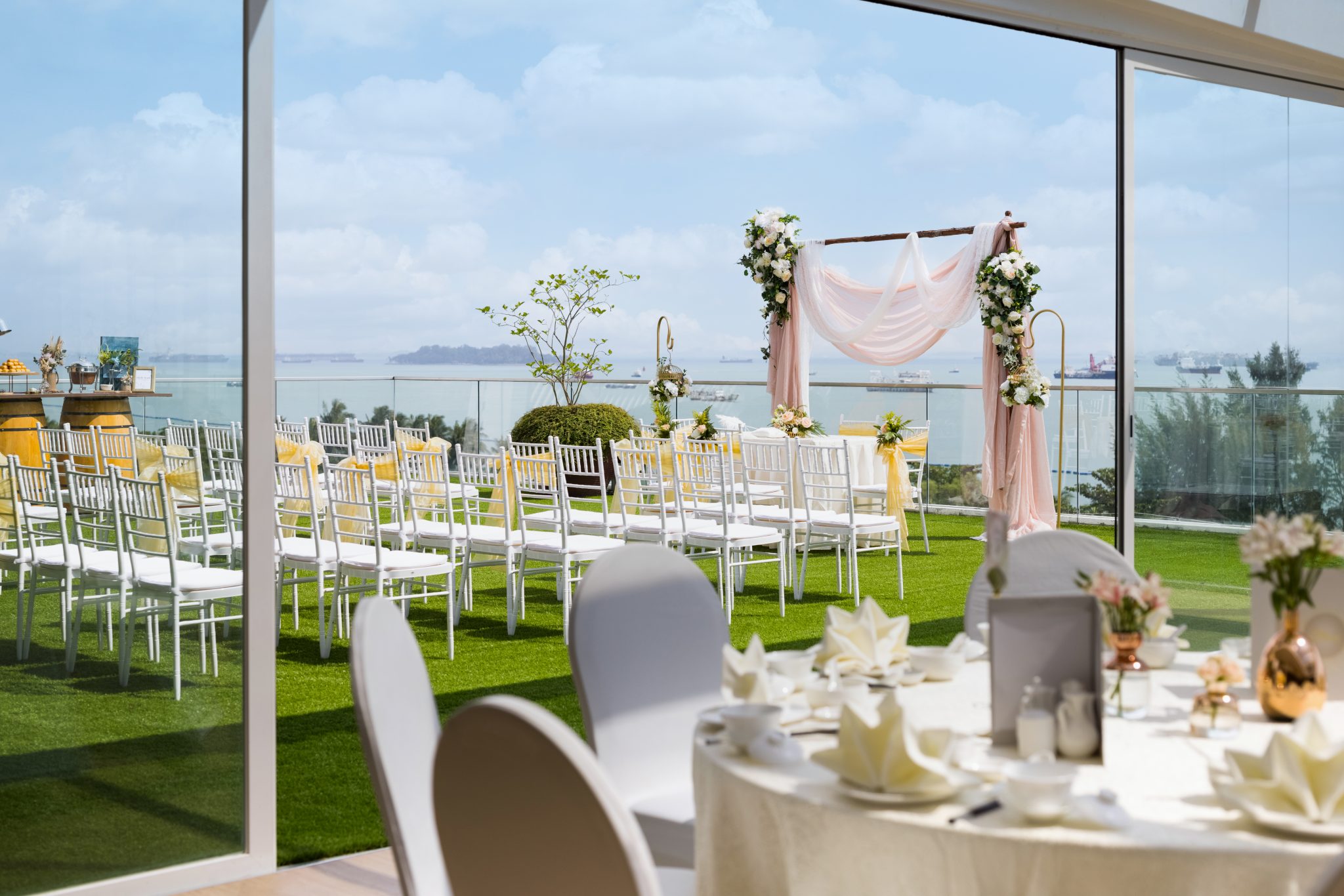 An outdoor rooftop wedding setup on green turf featuring white chairs, a pink fabric arch, and a panoramic ocean view.