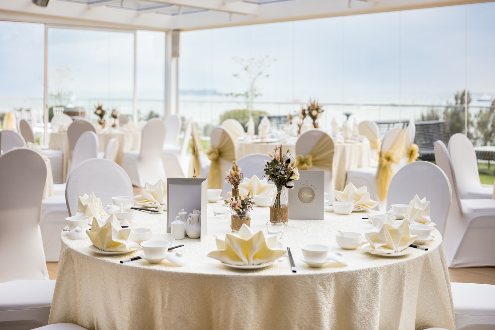 An elegant indoor wedding banquet setup with round tables, white linens, and floral centerpieces with a seaside view.