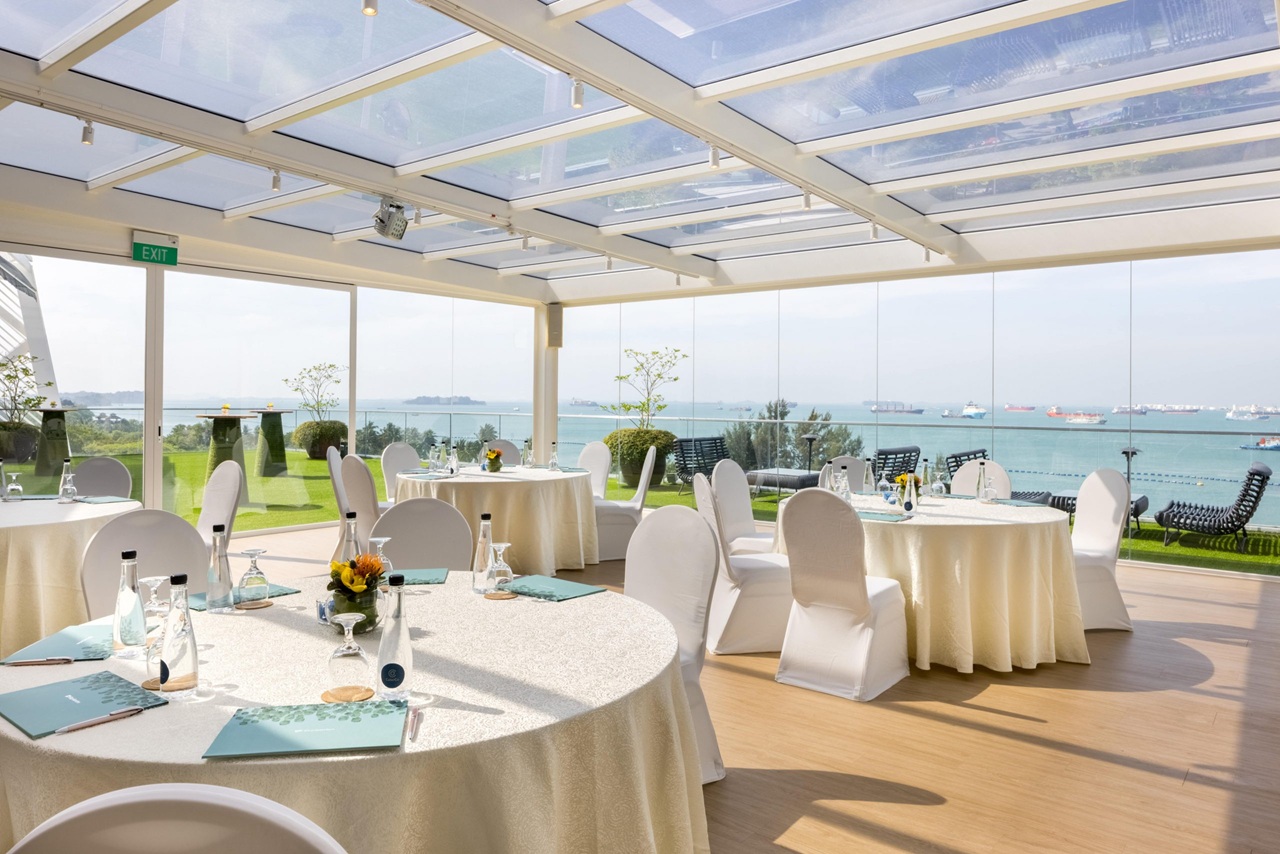 An indoor pavilion event space with round banquet tables, white chairs, a glass ceiling, and panoramic ocean views.