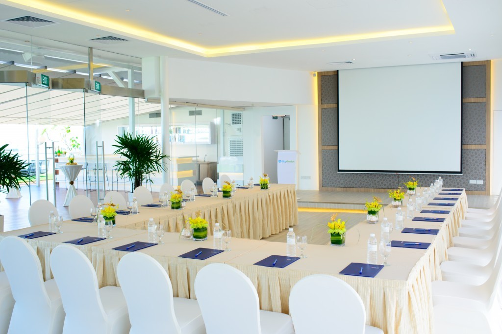 A corporate conference room setup with a U-shaped table, white chairs, floral centerpieces, and a projector screen.
