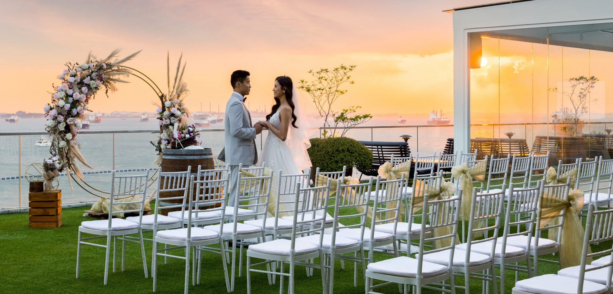 Bride and groom holding hands during a rooftop wedding ceremony at sunset, with an ocean backdrop and white guest chairs
Hyperlink