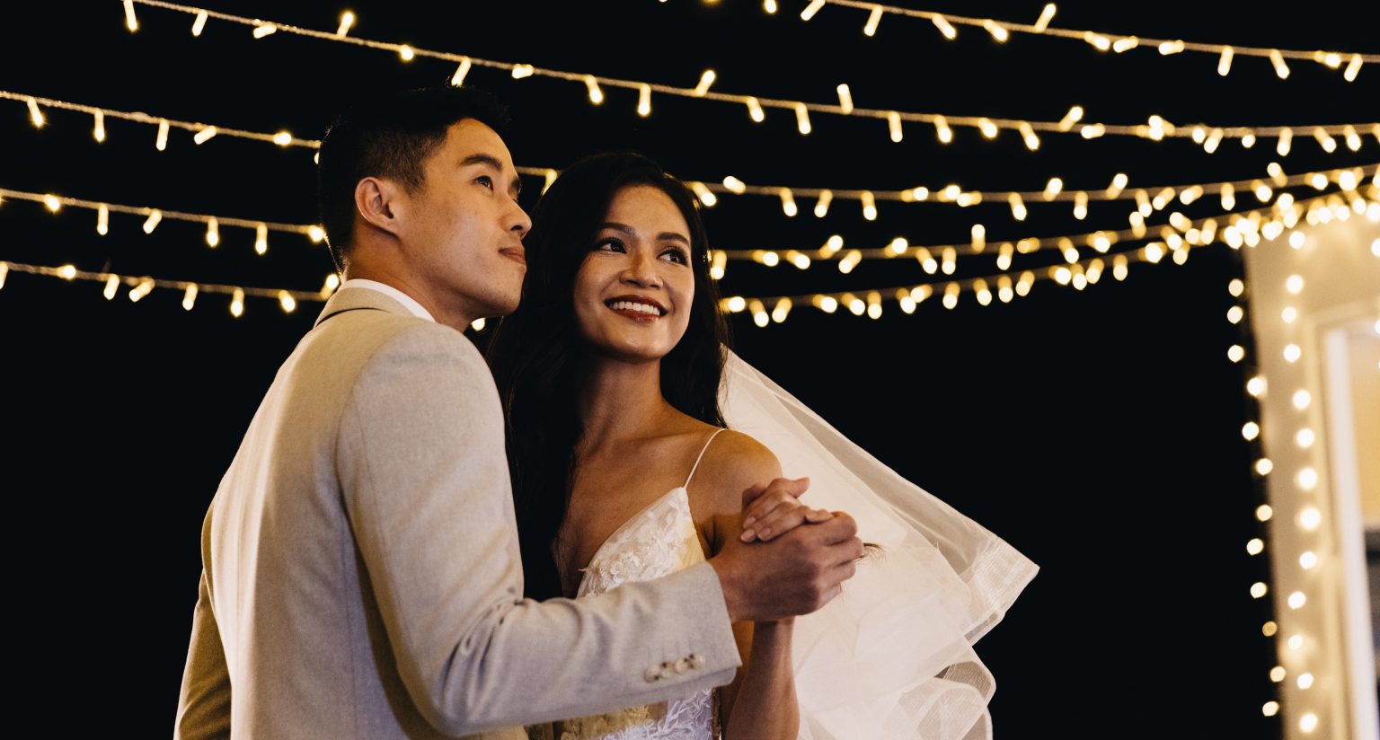 Smiling bride and groom holding hands and dancing during an outdoor night wedding reception lit by fairy lights.